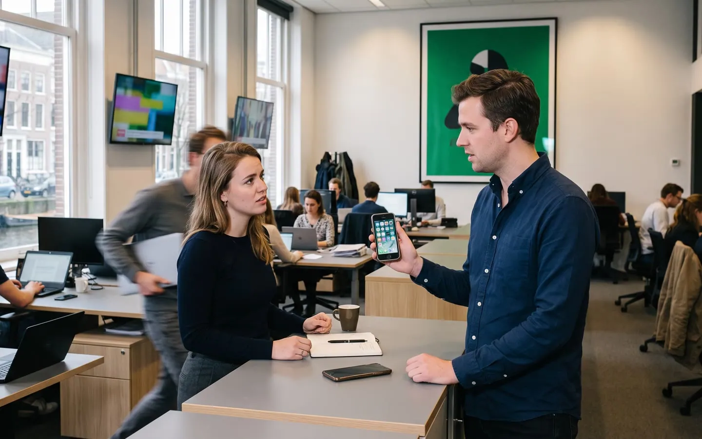 Een open redactievloer: twee redacteuren overleggen over een bericht op een telefoon, met schermen en bureaus erachter.